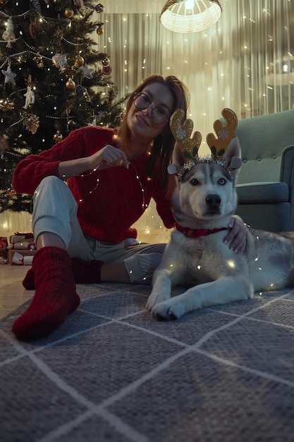 La giovane donna la sera di Natale insieme al cane si diverte a indossare le corna di cervo di Natale e la ghirlanda seduta sul pavimento davanti all'albero di Natale guarda la fotocamera