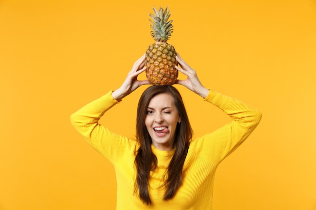 La giovane donna lampeggiante in abiti casual che mostra la lingua, tiene il frutto fresco dell'ananas maturo sulla testa isolata su sfondo giallo arancio. Persone stile di vita vivido, relax concetto di vacanza. Mock up copia spazio