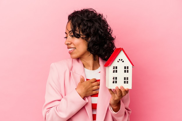 La giovane donna latina che tiene in mano una casa giocattolo isolata su sfondo rosa sembra da parte sorridente, allegra e piacevole.