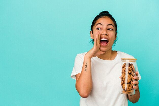 La giovane donna latina che tiene un barattolo di biscotti isolato su sfondo blu sta dicendo una notizia segreta di frenata calda e guarda da parte