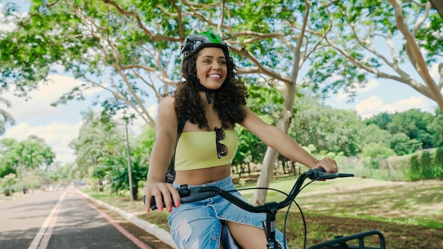 La giovane donna latina con il casco protettivo sta andando in bicicletta lungo la pista ciclabile in un parco cittadino