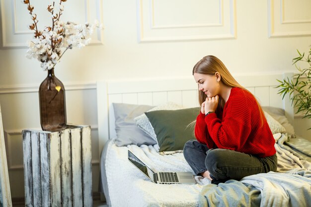 La giovane donna lavora al computer, seduta su un letto, lavorando a distanza. Una ragazza con i capelli lunghi in un maglione rosso e jeans lavora a casa.