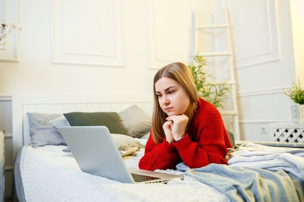 La giovane donna lavora al computer, seduta su un letto, lavorando a distanza. Una ragazza con i capelli lunghi in un maglione rosso e jeans lavora a casa.