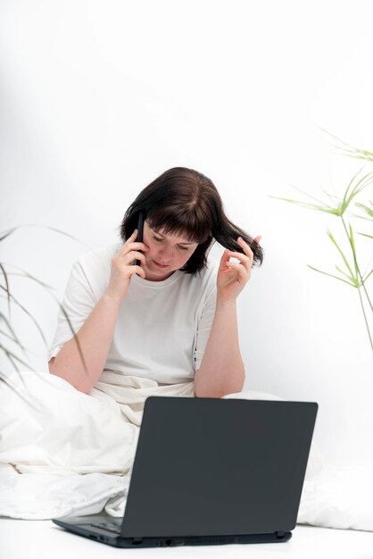 La giovane donna lavora seduta a letto. La libera professionista femminile lavora a letto e parla al telefono.