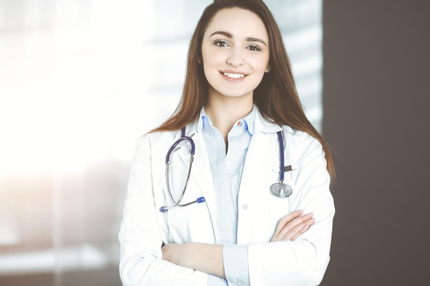 La giovane donna-medico sorridente è in piedi nella clinica soleggiata al chiuso.