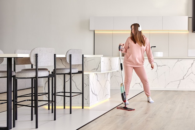 La giovane donna millenaria pulisce il pavimento in cucina ascoltando musica tramite cuffie wireless e ballando