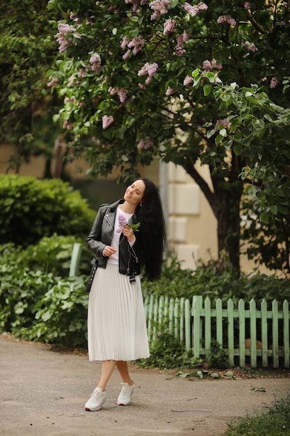 La giovane donna modello in gonna bianca e giacca di pelle nera si sta godendo un albero di lillà in fiore il giorno di primavera