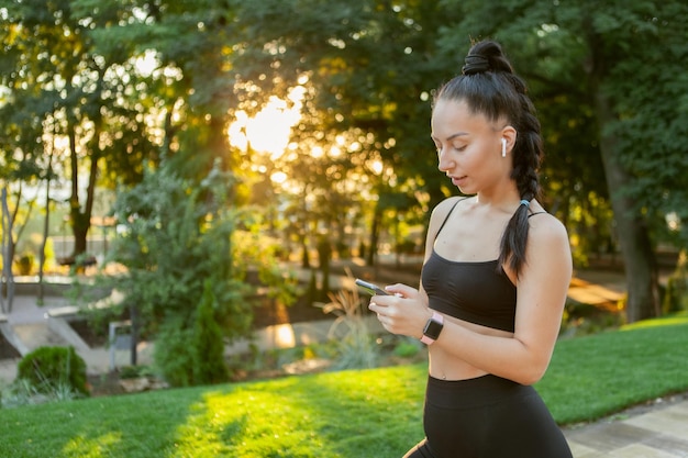 La giovane donna moderna di forma fisica in abiti sportivi ascolta la musica sulle cuffie e tiene un telefono nel parco all'alba