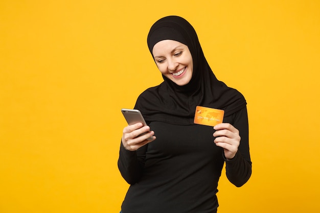 La giovane donna musulmana araba in abiti neri hijab tiene in mano il telefono cellulare, carta di credito isolata sul ritratto giallo della parete. Concetto di stile di vita religioso della gente.