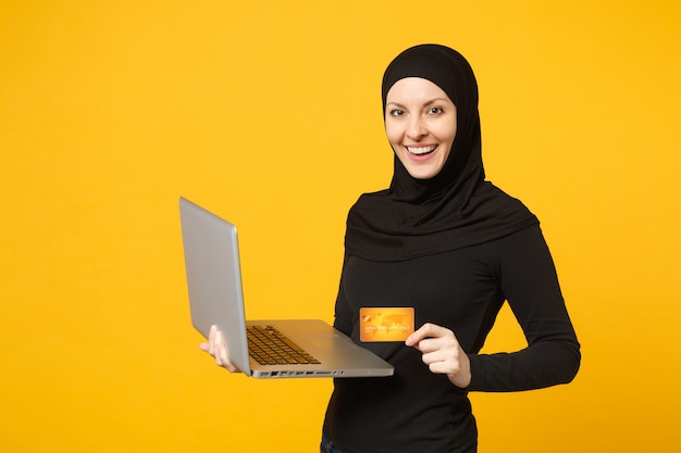 La giovane donna musulmana araba sorridente in vestiti neri di hijab tiene il pc del computer portatile, carta bancaria di credito isolata sul ritratto giallo della parete. Concetto di stile di vita religioso della gente.