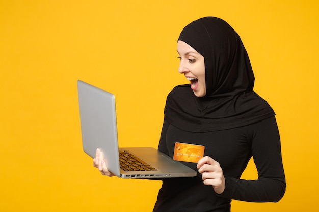 La giovane donna musulmana araba sorridente in vestiti neri di hijab tiene il pc del computer portatile, carta bancaria di credito isolata sul ritratto giallo della parete. Concetto di stile di vita religioso della gente.