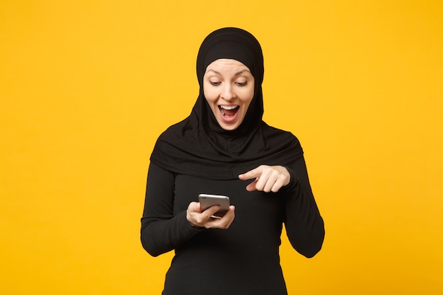 La giovane donna musulmana araba sorridente in vestiti neri di hijab tiene in mano il cellulare, chiacchierando isolato sul ritratto giallo della parete. Concetto di stile di vita religioso della gente. .