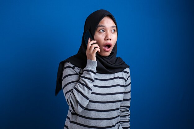 La giovane donna musulmana asiatica che indossa l'hijab mostra un'espressione scioccata mentre riceve una telefonata su sfondo blu