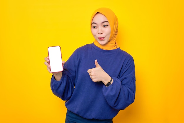 La giovane donna musulmana asiatica eccitata vestita con un maglione casual che mostra il telefono cellulare con schermo vuoto fa il gesto del pollice su raccomandando un'app mobile isolata su sfondo giallo