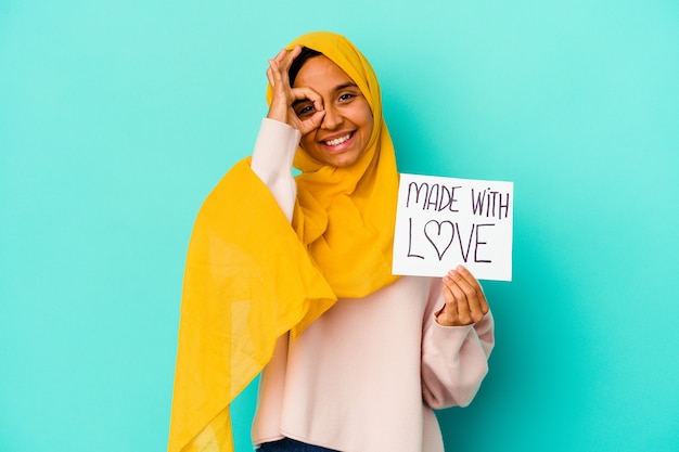 La giovane donna musulmana che tiene un fatto con il cartello di amore isolato sulla parete blu ha eccitato mantenendo il gesto giusto sull'occhio.