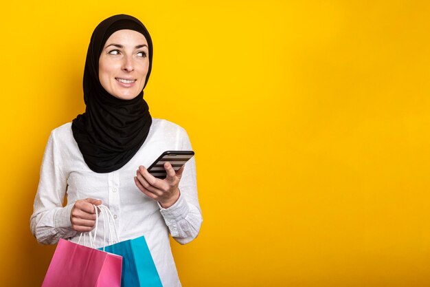 La giovane donna musulmana in hijab tiene un telefono e borse della spesa su sfondo giallo Banner