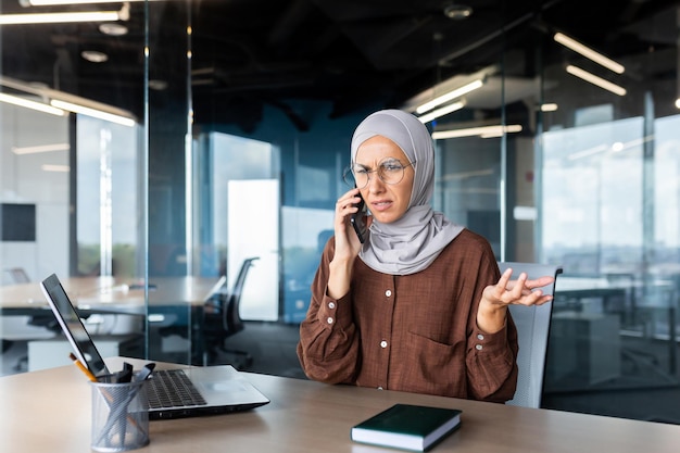 La giovane donna musulmana preoccupata in hijab seduta in ufficio e che parla al telefono risolve le domande dei problemi