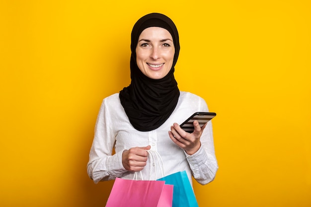 La giovane donna musulmana sveglia in hijab tiene il telefono e le borse della spesa su colore giallo