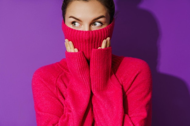 La giovane donna nasconde il viso all'interno di un maglione a collo alto caldo e accogliente su sfondo viola