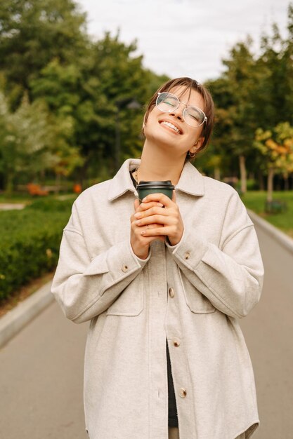 La giovane donna nel parco tiene stretto e gode di una tazza di caffè caldo per andare