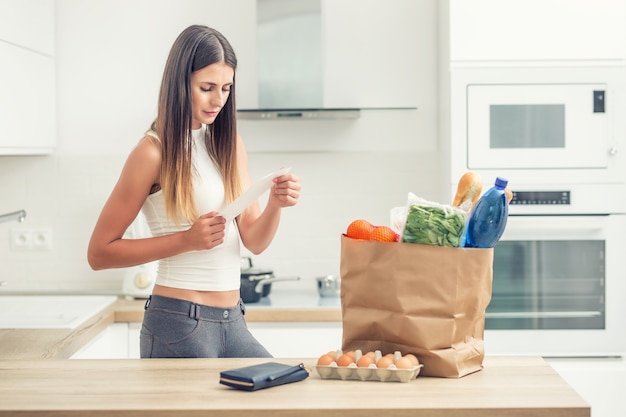 La giovane donna nella cucina domestica controlla il disegno di legge. Acquista su un tavolo in un sacchetto di carta.