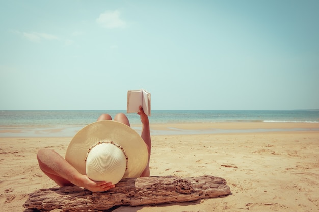 La giovane donna nella menzogne del cappello di paglia prende il sole su una spiaggia tropicale, si rilassa con il libro.