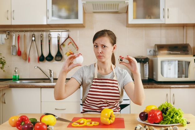 La giovane donna pensierosa attraente in un grembiule sceglie tra pollo e uova di quaglia in cucina. Concetto di dieta. Uno stile di vita sano. Cucinare a casa. Prepara da mangiare.