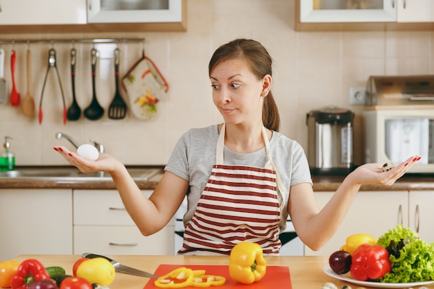 La giovane donna pensierosa attraente in un grembiule sceglie tra pollo e uova di quaglia in cucina. Concetto di dieta. Uno stile di vita sano. Cucinare a casa. Prepara da mangiare.