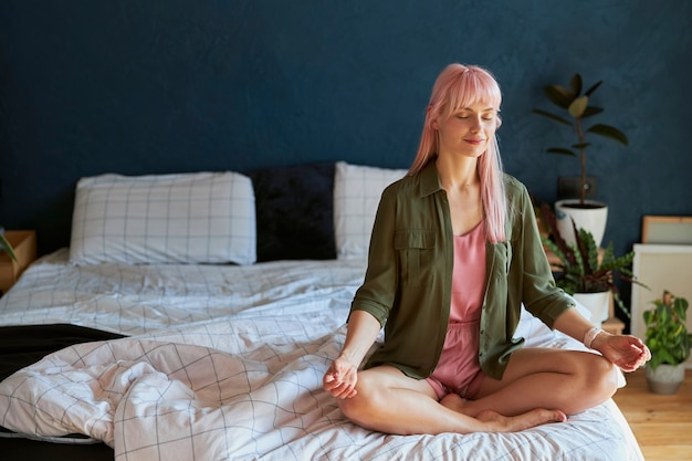 La giovane donna positiva in camicia verde e pigiama medita seduta nella posa del loto su un letto grande