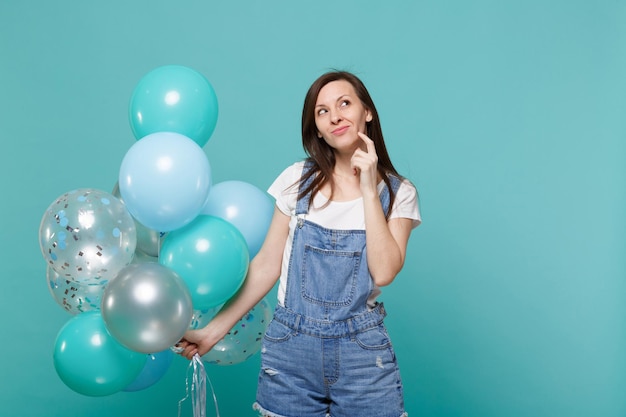 La giovane donna premurosa in abiti in denim che guarda in alto ha messo la mano sul mento per celebrare la tenuta di palloncini colorati isolati su sfondo blu turchese. Festa di compleanno, concetto di emozioni della gente.