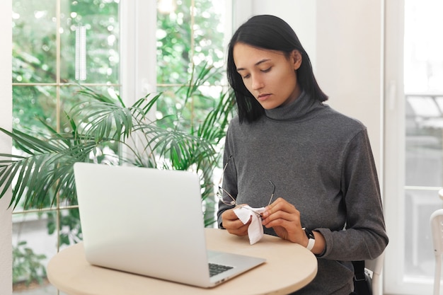 La giovane donna pulisce gli occhiali mentre lavora al computer portatile