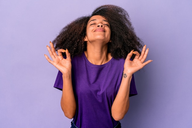 La giovane donna riccia afroamericana isolata sulla parete viola si rilassa dopo una dura giornata di lavoro, sta eseguendo yoga.