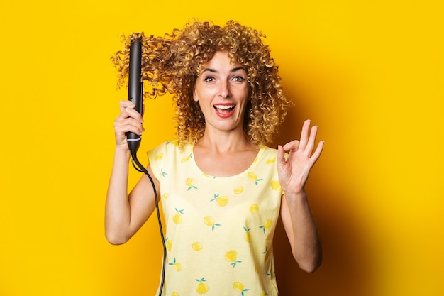 La giovane donna riccia allegra raddrizza la piastra per capelli su sfondo giallo.