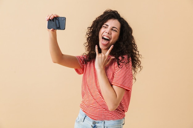 La giovane donna riccia emotiva fa selfie che mostra il gesto della roccia.