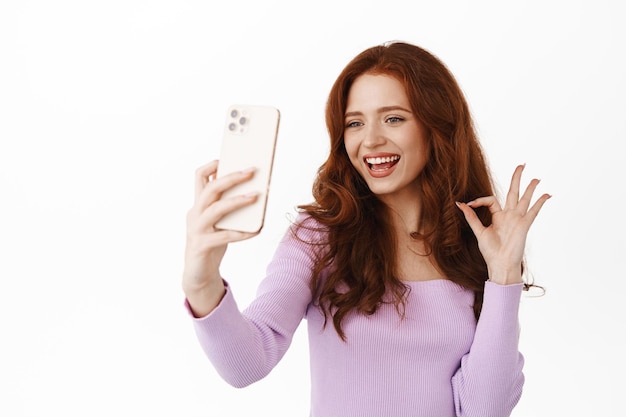 La giovane donna rossa sorridente mostra il segno giusto durante la videochiamata sullo smartphone, approva o elogia qualcosa di buono, scatta un selfie, registra un videomessaggio, sfondo bianco