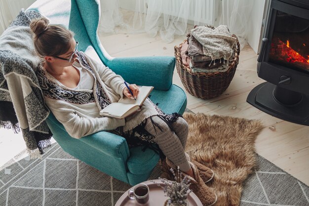 La giovane donna scrive su un taccuino seduta su un'accogliente poltrona accanto al caminetto