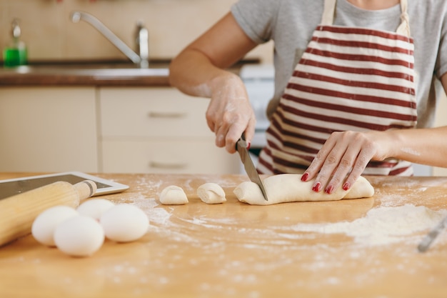 La giovane donna seduta a un tavolo con tavoletta, taglia a pezzi un impasto con un coltello e va a preparare una torta in cucina. Cucinare a casa. Preparare il cibo da vicino.