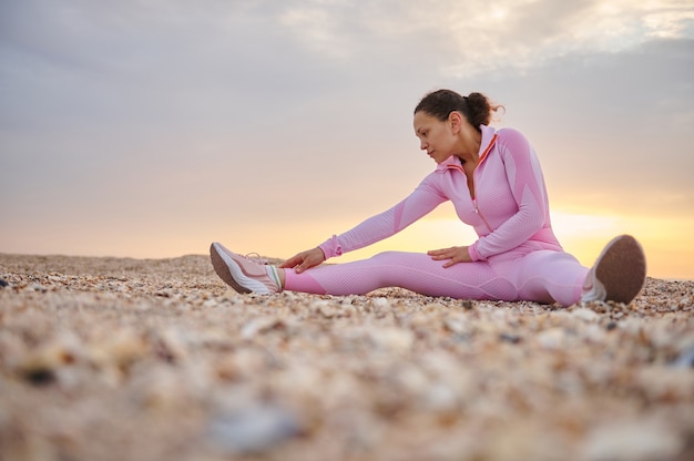 La giovane donna seduta sulla spiaggia si è concentrata sull'allungamento del corpo durante l'allenamento mattutino all'alba