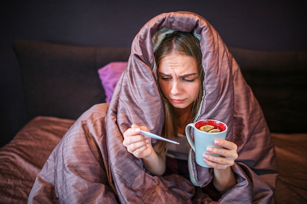 La giovane donna seria ma malata si siede sul letto con la coperta coperta. Tiene in mano una tazza di tè caldo e un termometro. La ragazza lo guarda. Lei soffre.