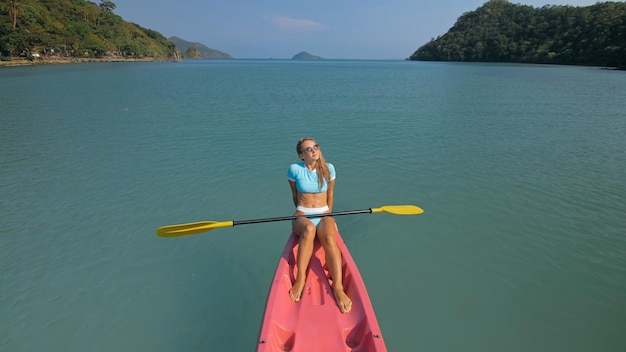 La giovane donna sexy in costume da bagno blu oscilla i piedi in acqua sulla canoa rosa sul mare contro la parte posteriore delle colline