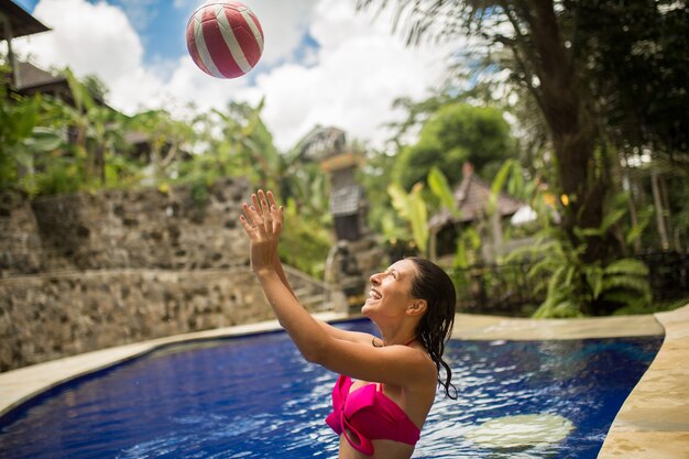La giovane donna sexy in costume da bagno rosa gioca con la palla nella piscina tropicale.
