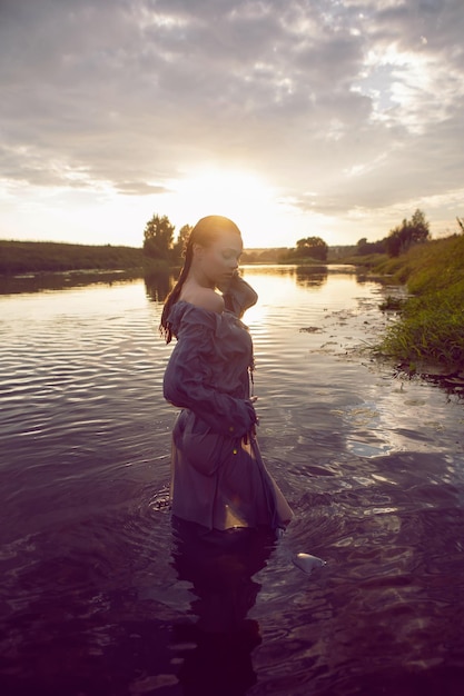 La giovane donna sexy in un vestito grigio sta nell'acqua al tramonto nel fiume