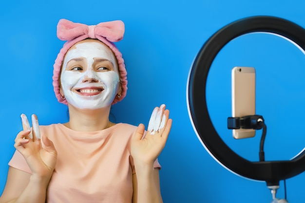La giovane donna si applica una maschera cosmetica di crema sul viso e comunica con i suoi amici online sul suo smartphone