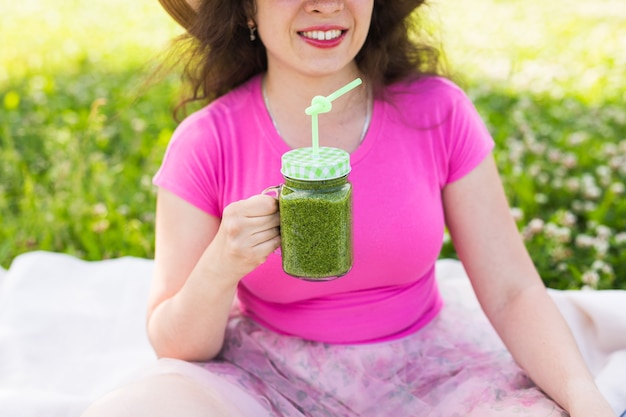 La giovane donna si diverte nel parco e beve frullati verdi durante un picnic.