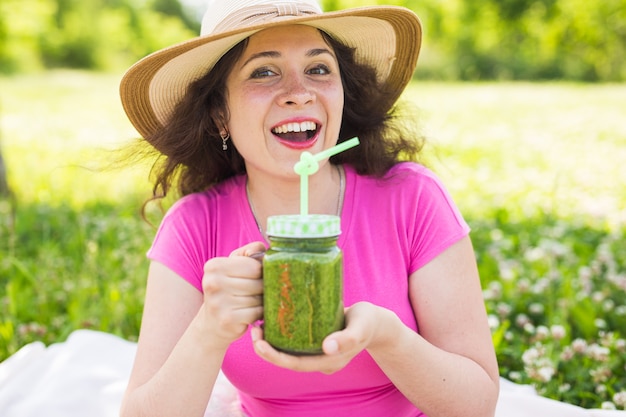 La giovane donna si diverte nel parco e beve frullati verdi durante un picnic.