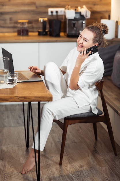 La giovane donna si siede al tavolo della cucina utilizzando un computer portatile e parlando su un telefono cellulare e sorridente. Riuscita ragazza che ride e che lavora a casa. Bella donna alla moda che sorride e che si rilassa a casa.