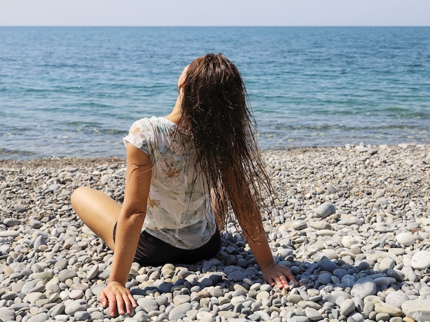 La giovane donna si siede con la schiena sulla spiaggia di ciottoli e scuote i capelli bagnati