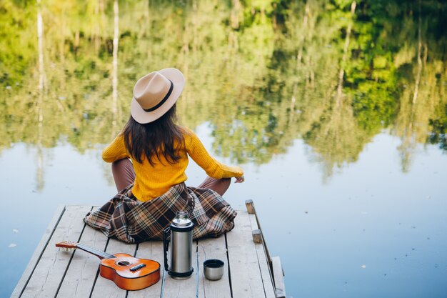 La giovane donna si siede con una chitarra su un ponte su un lago con un paesaggio autunnale. Tonificante.