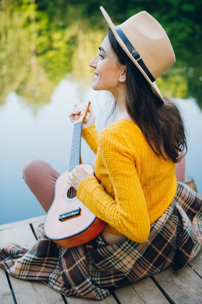 La giovane donna si siede con una chitarra su un ponte su un lago con un paesaggio autunnale. Tonificante.