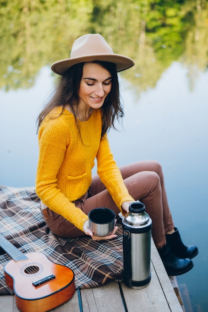 La giovane donna si siede su un ponte su un lago con un paesaggio autunnale e beve tè caldo da un thermos. Tonificante.